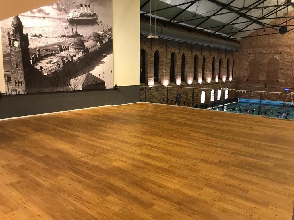 Leere Trainingsfläche mit durchgehendem Boden in heller Holzoptik in einer historischen Halle; links ein großformatiges Schwarz-Weiß-Foto des Hamburger Hafens, rechts hinter einem Geländer ein Schwimmbecken unter einem Sheddach.