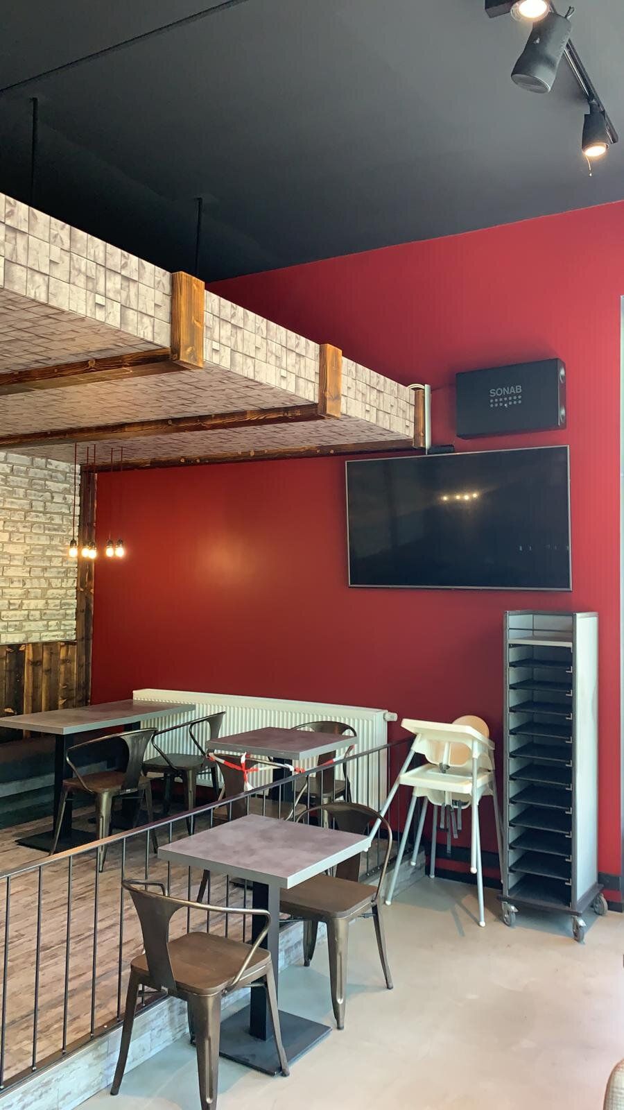 Sitzbereich einer Bäckereifiliale auf zwei Ebenen: rote Wand mit Bildschirm und Lautsprecher, Bistrotische mit Metallstühlen, Galeriekante mit Stahlgeländer und rot-weißem Absperrband, darüber eine Verkleidung in Ziegeloptik mit Holzbalken.