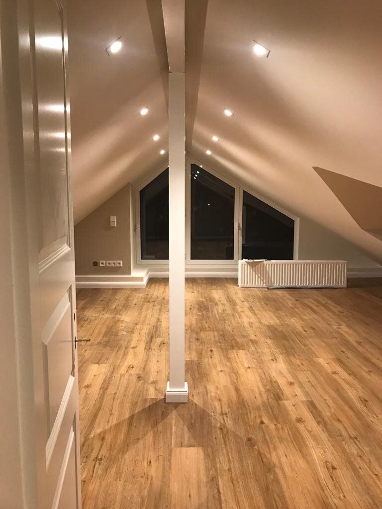 Ausgebautes Dachgeschoss am Abend: heller Boden in Holzoptik, Dachschrägen zu beiden Seiten, eine weiß verkleidete Stütze in der Raummitte, eine Fensterfront über einem Heizkörper, Einbaustrahler in der Schräge.