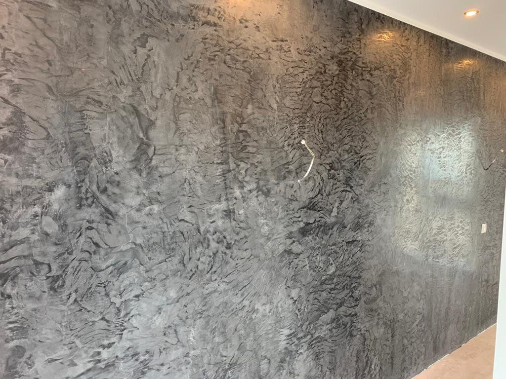 Wand mit dunkelgrauer, von Hand aufgezogener Spachtelstruktur im Streiflicht zweier Deckenstrahler; aus der Wand ragt ein weißes Kabelende für eine Wandleuchte.